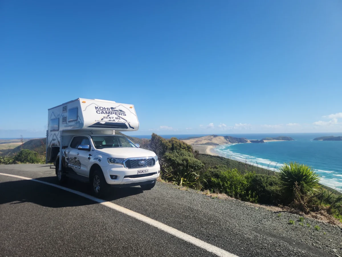 A white camper van is parked on a coastal road overlooking a scenic beach, blue ocean, and distant sand dunes under a clear blue sky.