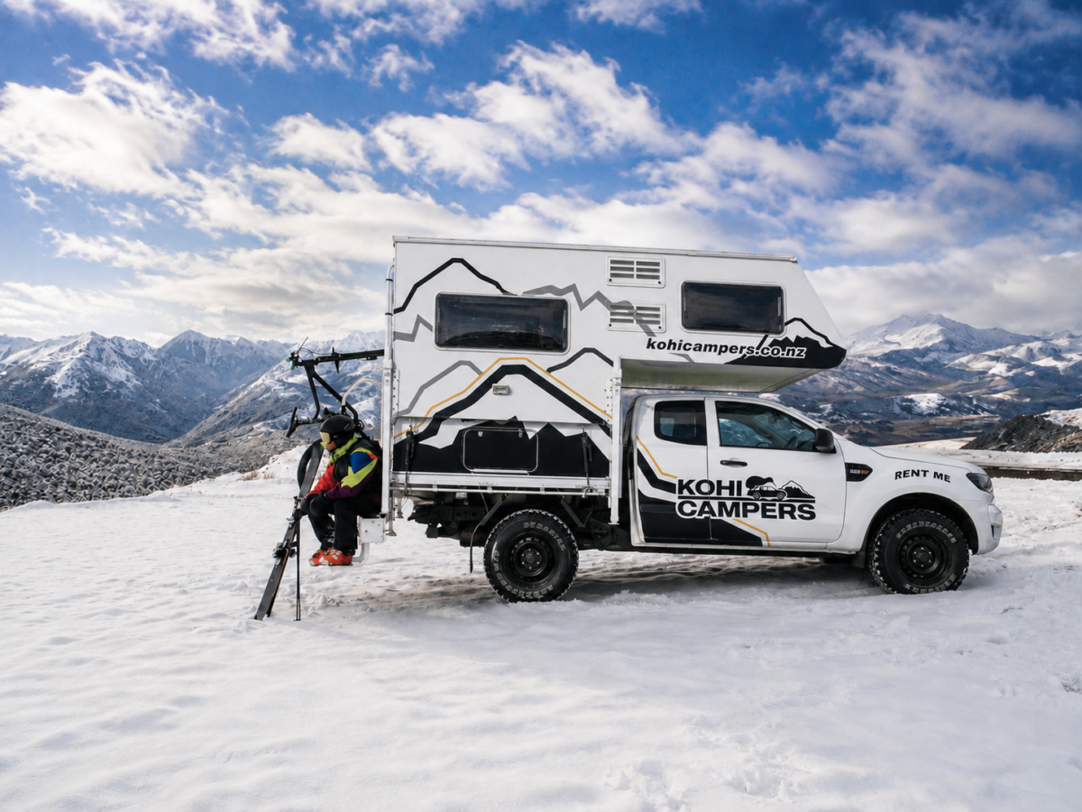 A person in ski gear sits on the back of a white Kohi Campers truck camper parked on snowy terrain, with mountains and a partly cloudy sky in the background.