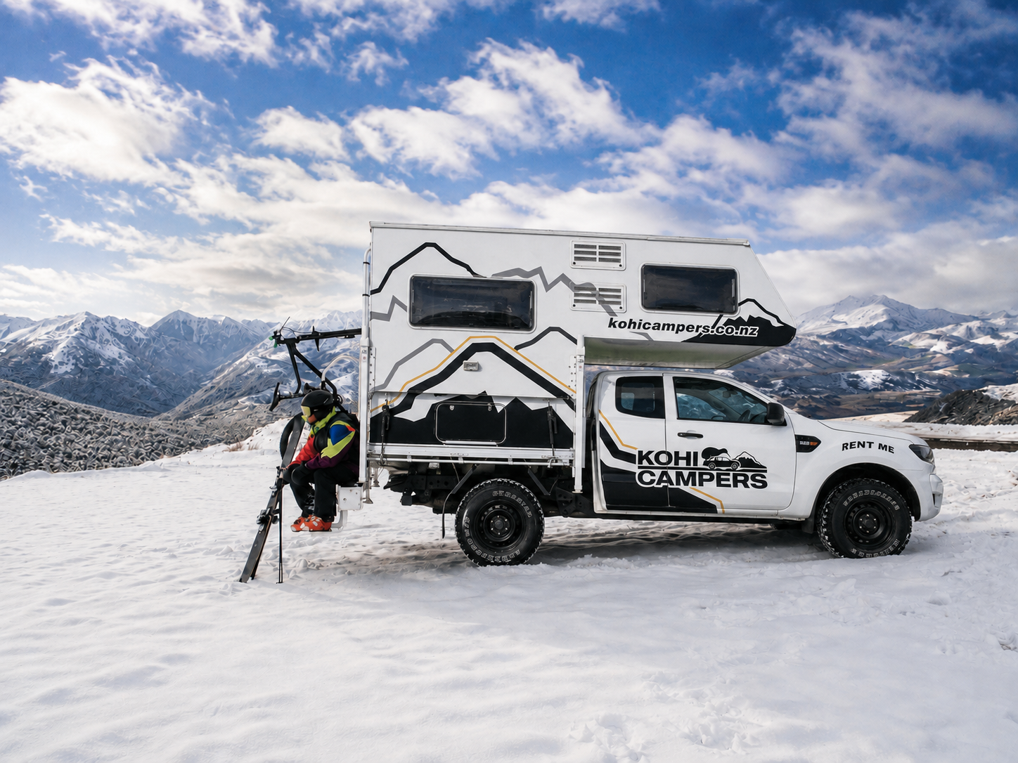 A person in ski gear sits on the back of a white Kohi Campers truck camper parked on snowy terrain, with mountains and a partly cloudy sky in the background.