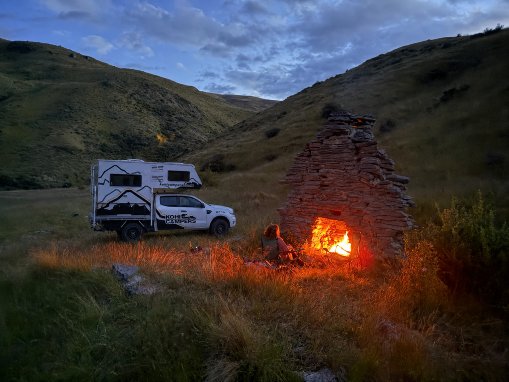 A person sits by a glowing campfire in a stone outdoor fireplace, beside a camper truck parked on grassy hills at dusk, with rolling hills and a cloudy sky in the background.