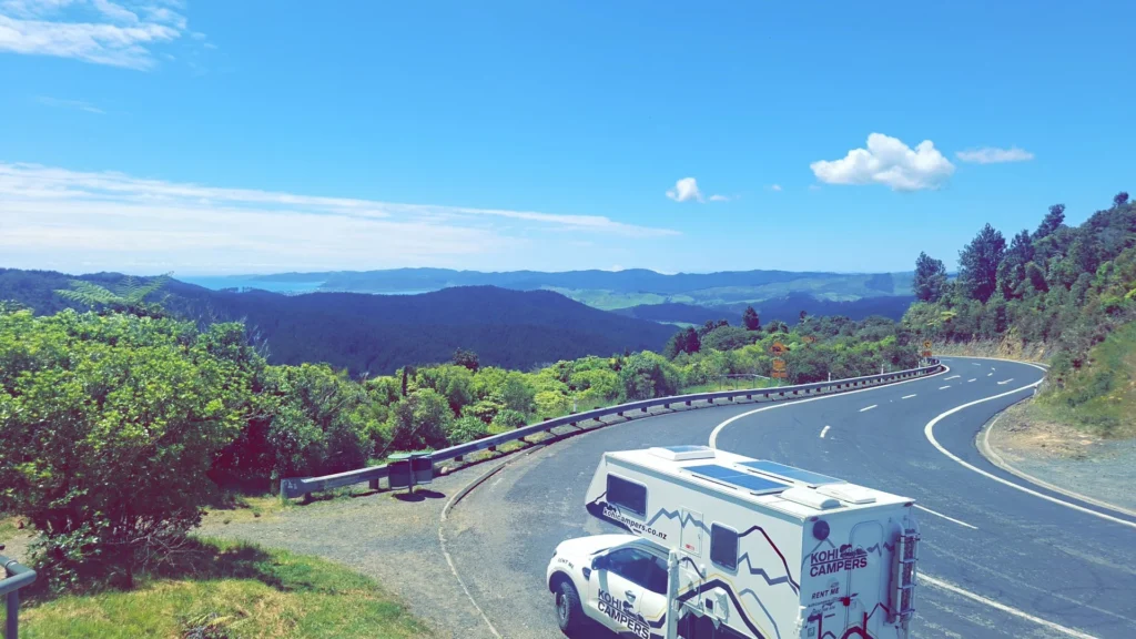 A white camper van parked on the side of a winding mountain road, surrounded by lush green trees and hills under a bright blue sky with scattered clouds.
