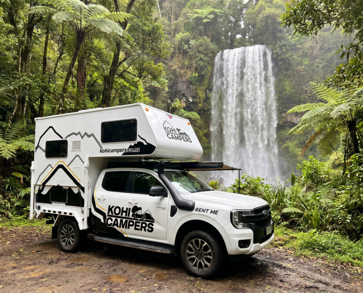 A white Kohi Campers rental camper truck is parked on a forested path near a large, cascading waterfall surrounded by lush green trees and foliage.