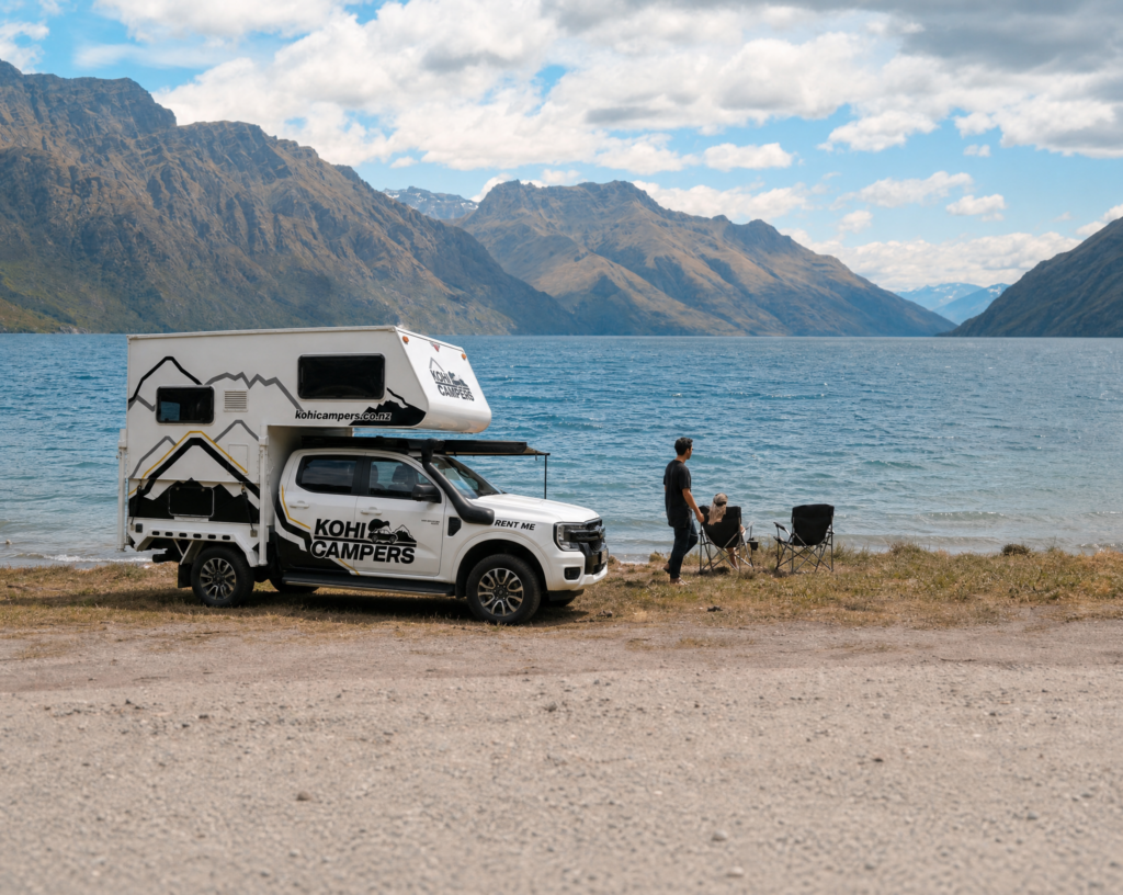 A white camper van parked beside a lake with mountains in the background. Two people relax on camping chairs near the water under a partly cloudy sky, enjoying the scenic landscape.