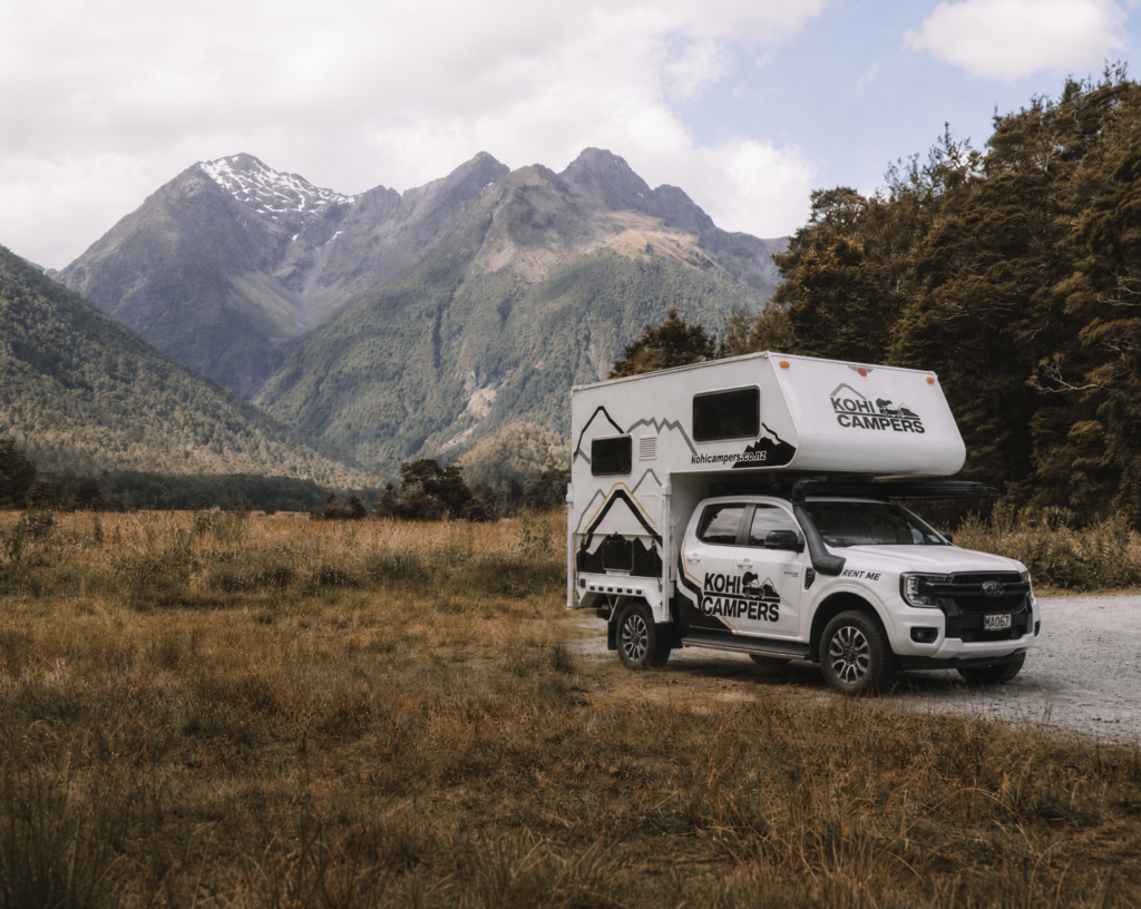 A white Kohi Campers RV is parked on a grassy field with tall mountains and a forest in the background under a cloudy sky.