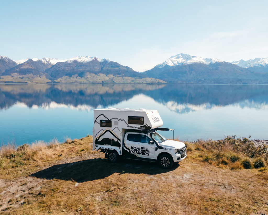 A white camper truck parked on grassy terrain beside a calm lake, with mountains and snow-capped peaks reflected in the water under a clear blue sky.