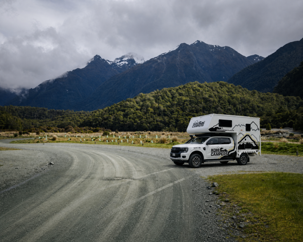A campervan parked on a gravel road curves through a scenic landscape with forested hills and snow-capped mountains under a cloudy sky.