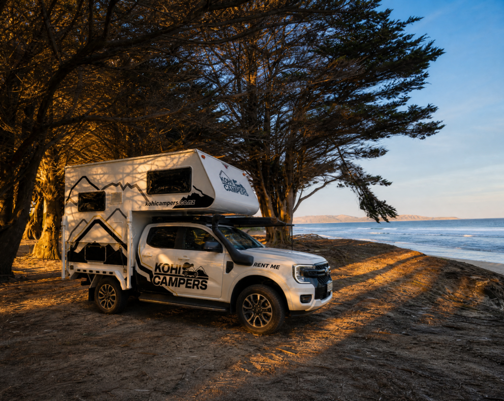 A white Kohi Campers truck with a mounted camper is parked under tall trees by the beach at sunset, with the ocean and distant hills visible in the background.