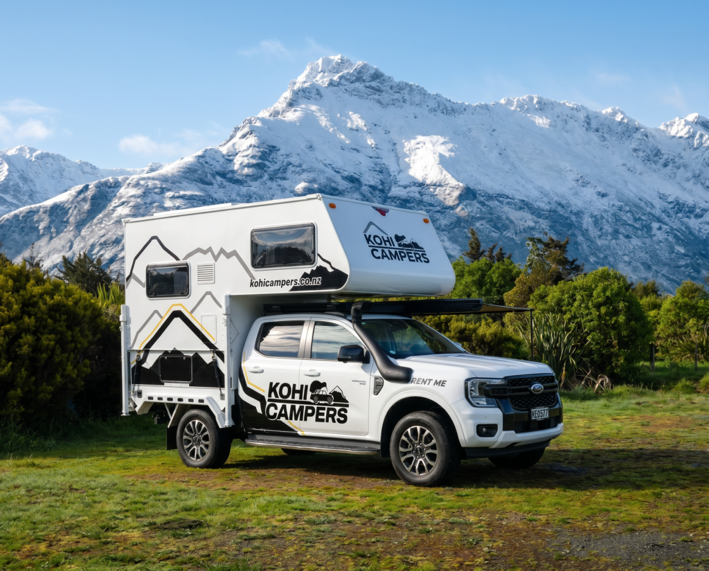 A white Kohi Campers rental campervan is parked on grass with snow-capped mountains and lush green bushes in the background under a clear blue sky.