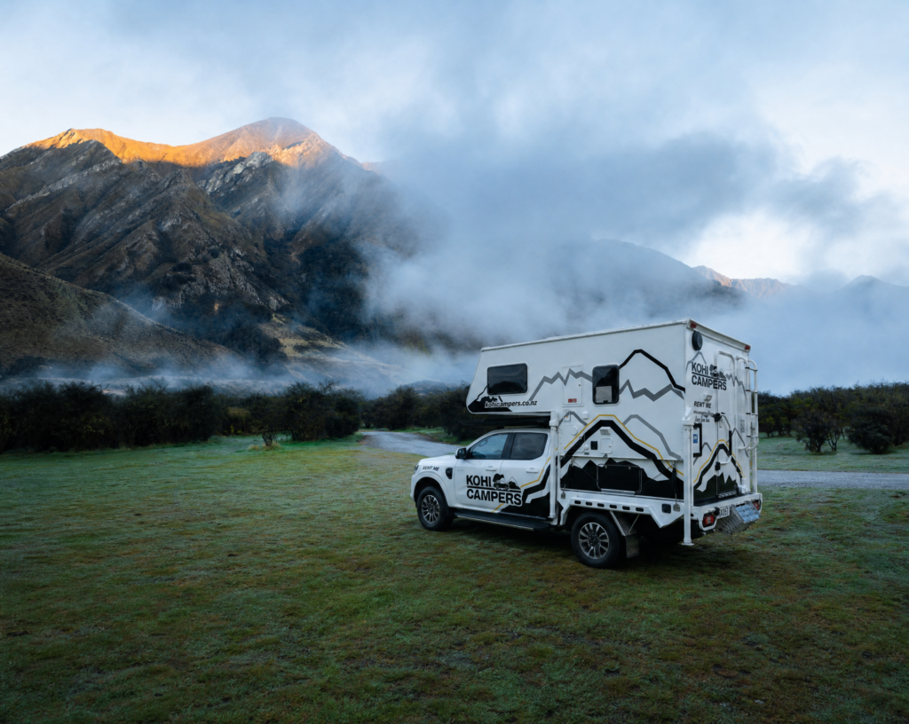 A campervan parked on a grassy field with misty mountains in the background at sunrise. The campervan has mountain graphics and “Kohu Campers” branding on its side. Low clouds partially cover the mountains.