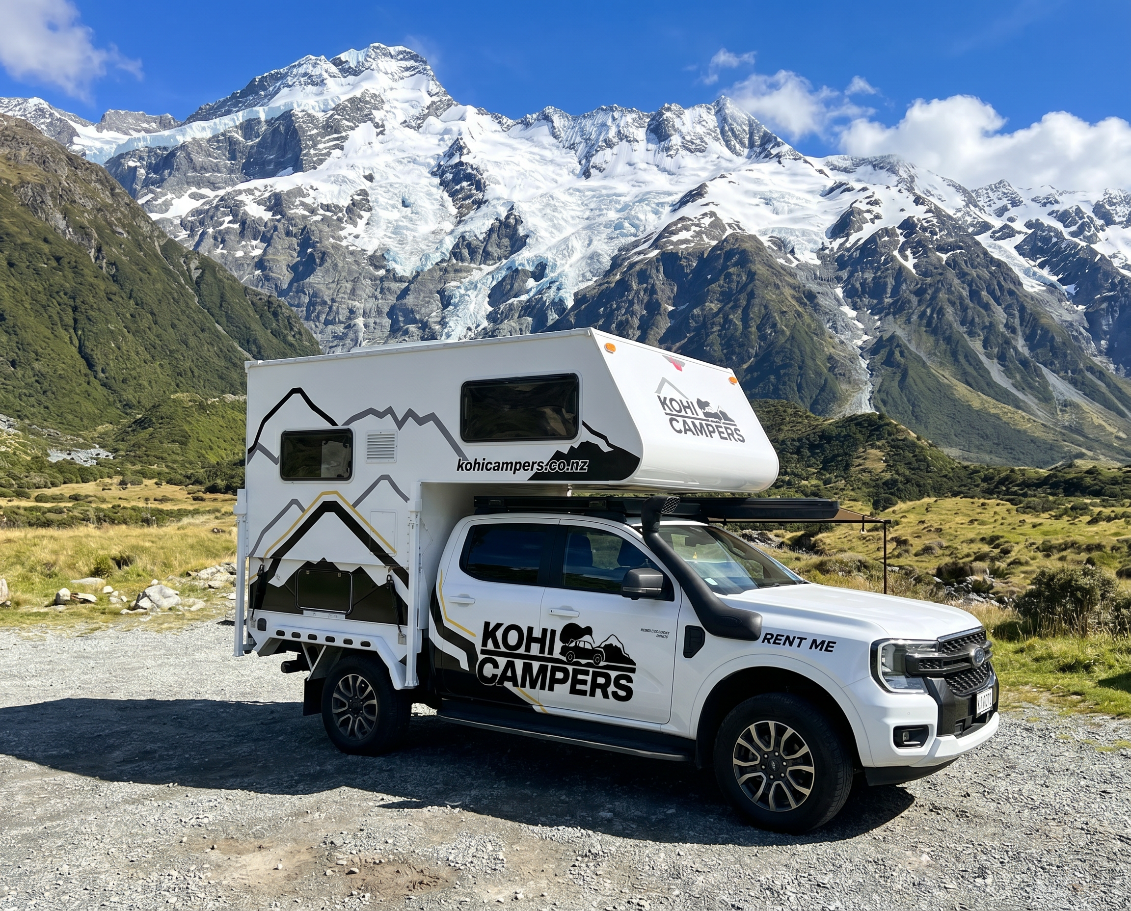 A white Kohi Campers rental camper truck is parked on a gravel road in a scenic mountain valley, with snow-capped peaks and blue sky in the background.