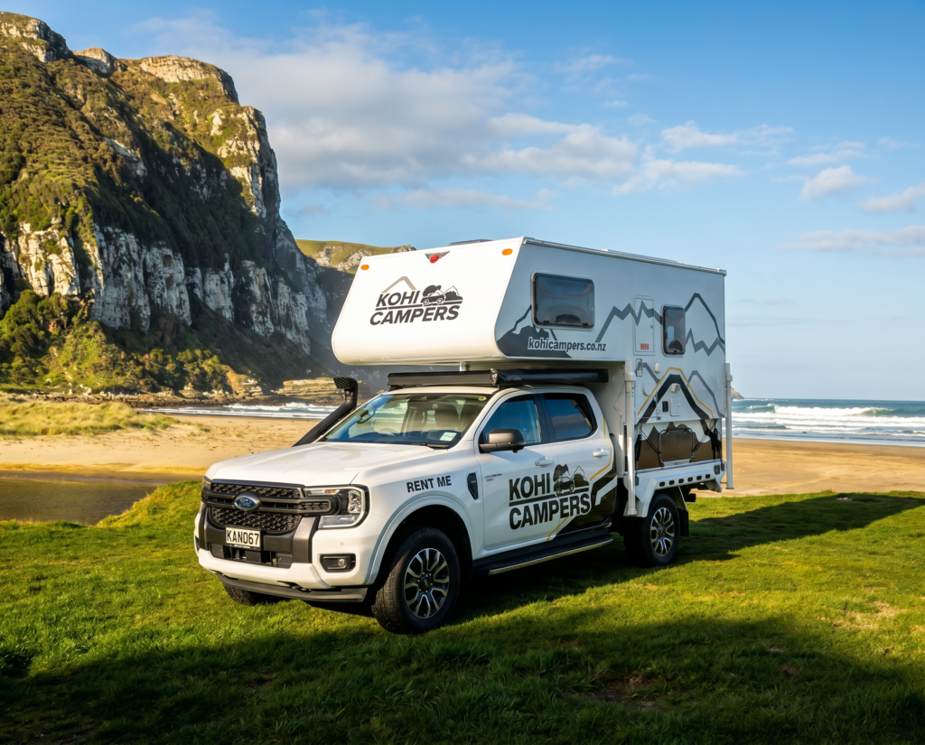 A white KOHI CAMPERS campervan is parked on green grass near a sandy beach, with rocky cliffs and blue sky in the background.
