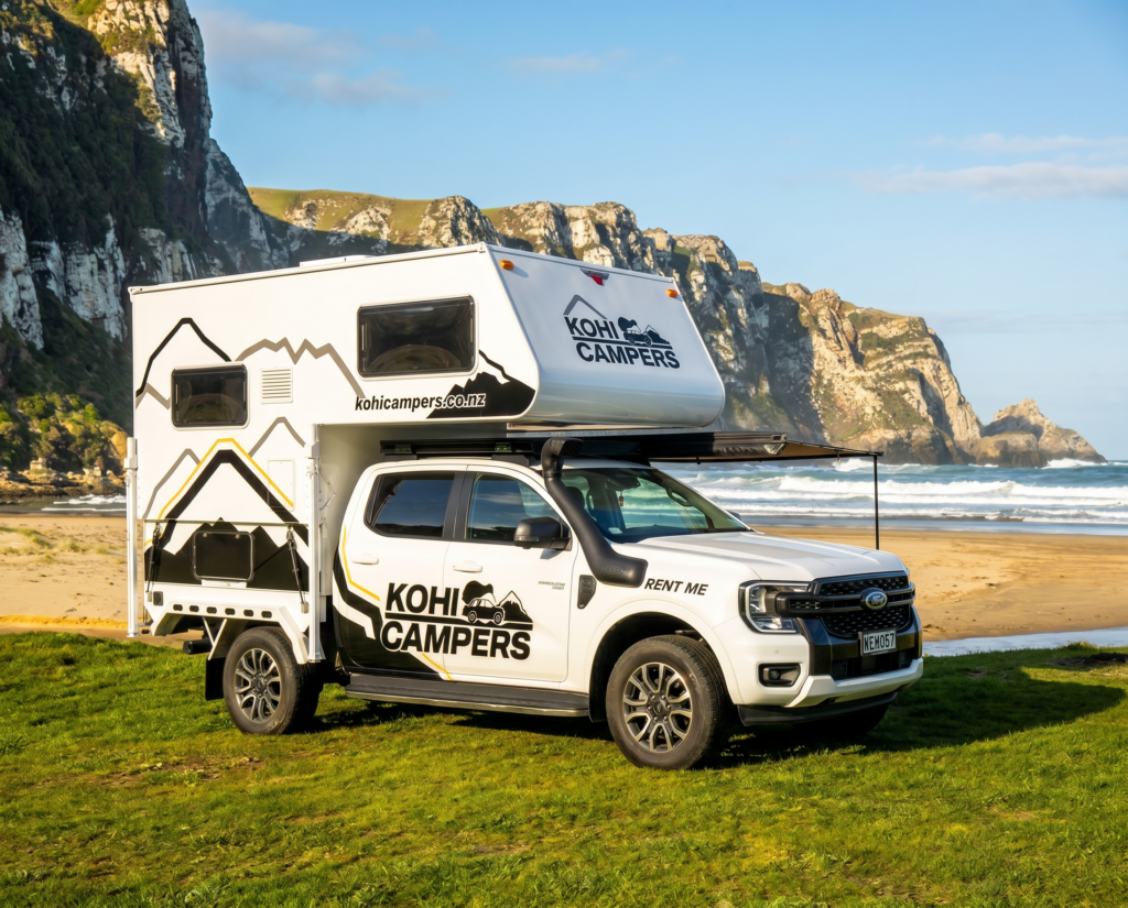 A white Kohi Campers RV is parked on grass near a sandy beach, with cliffs and blue sky in the background. The vehicle has RENT ME and mountain graphics on its side.