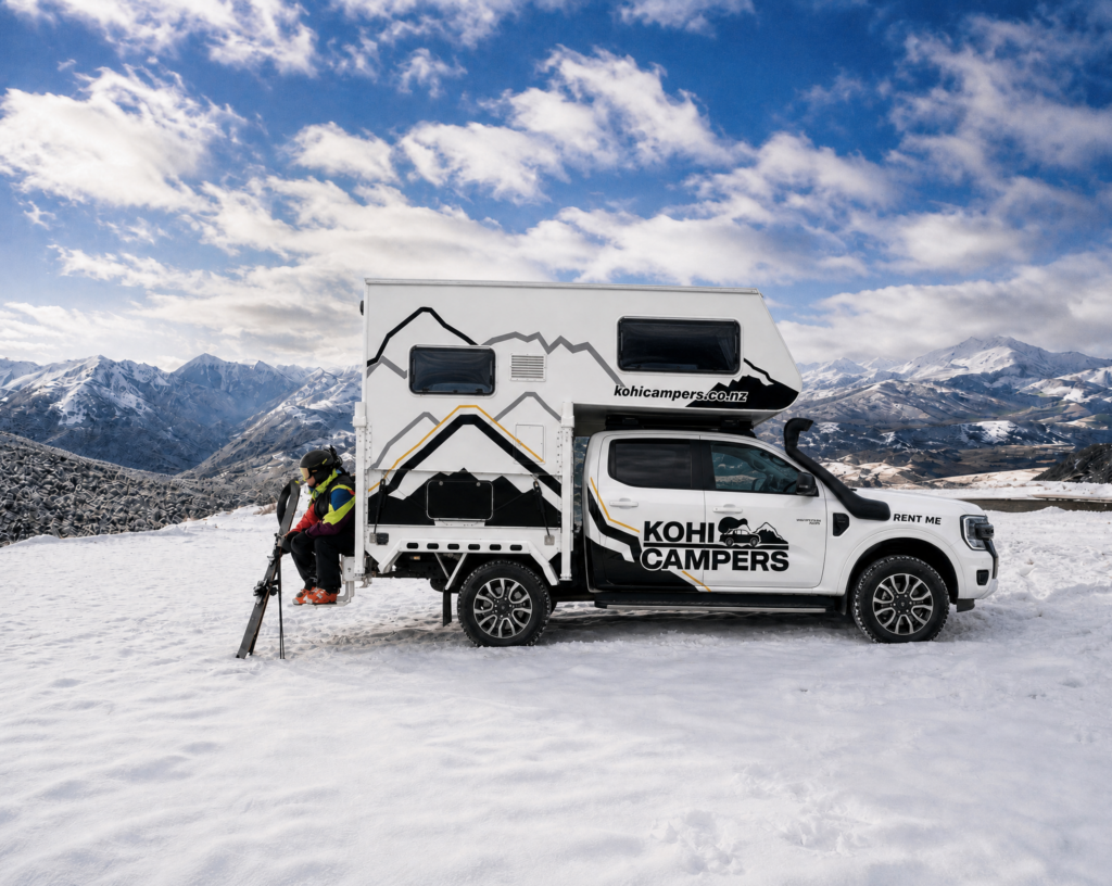 A person in winter gear and skis stands next to a white Kohi Campers truck camper parked on snow, with snowy mountains and a cloudy sky in the background.